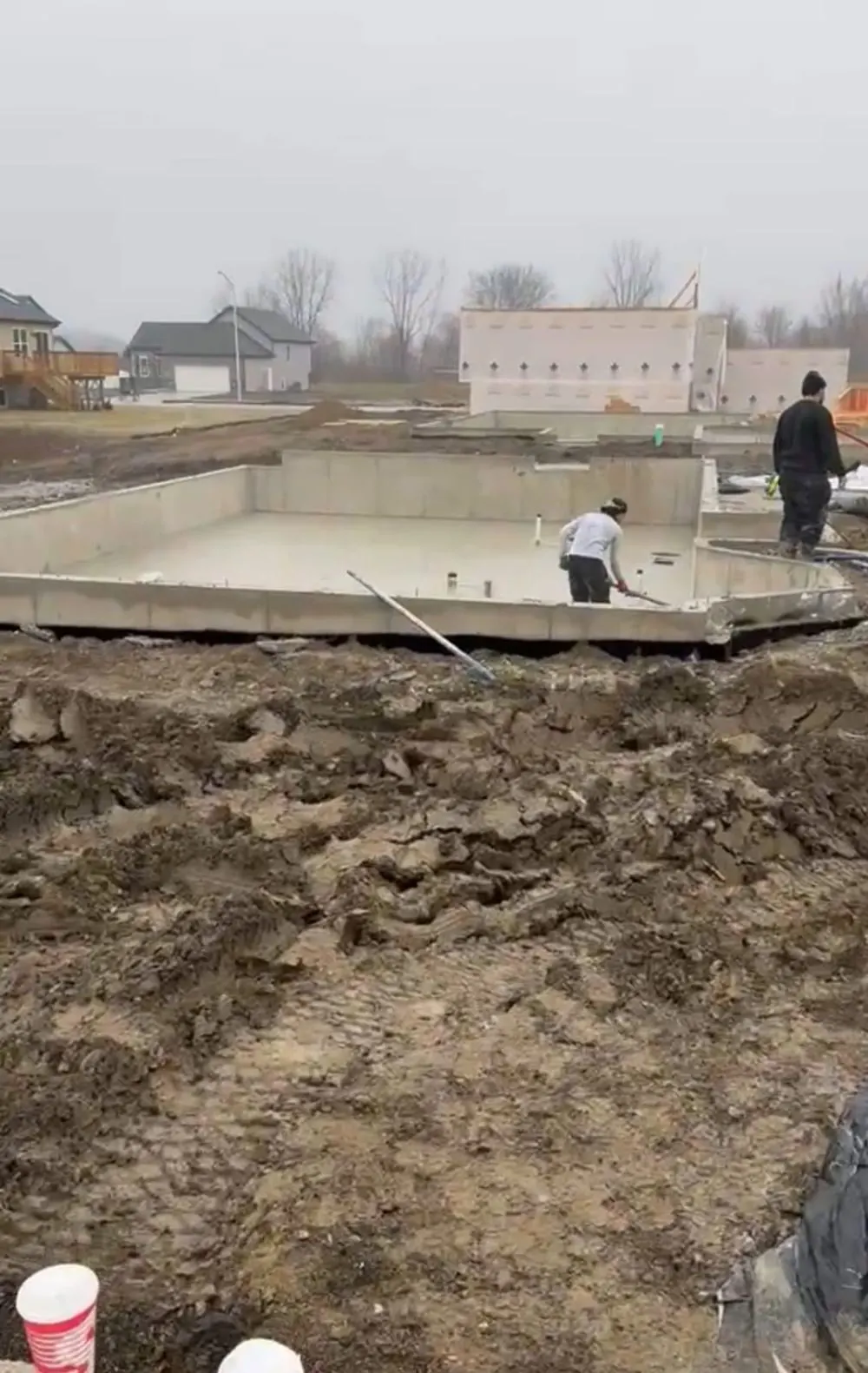 Crew finishing a poured basement slab inside foundation walls on a rainy job site, muddy excavation in the foreground