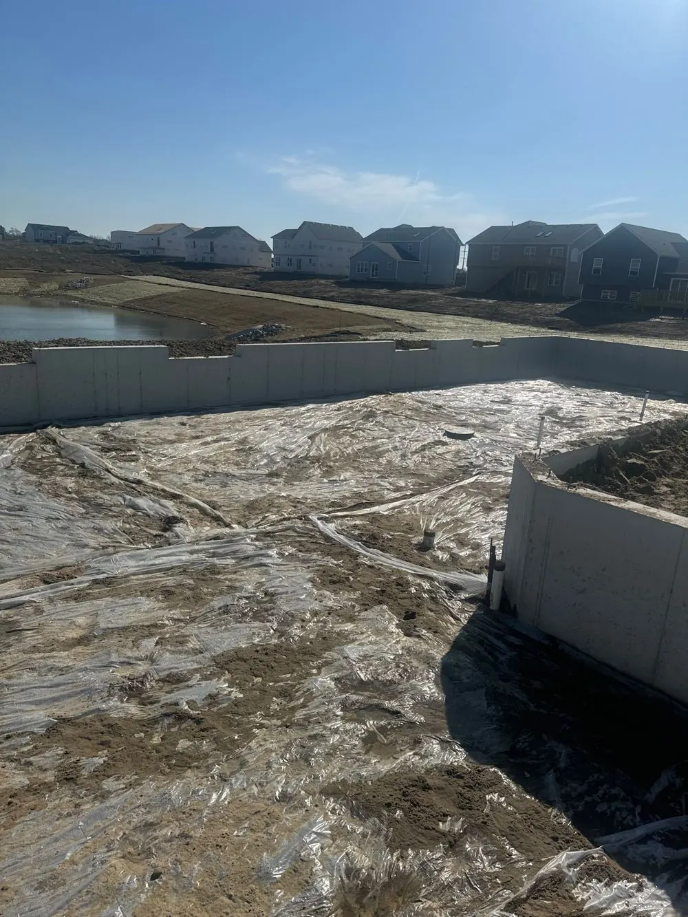 Basement slab job site covered in plastic vapor barrier before the pour, foundation walls up, pond and homes in background