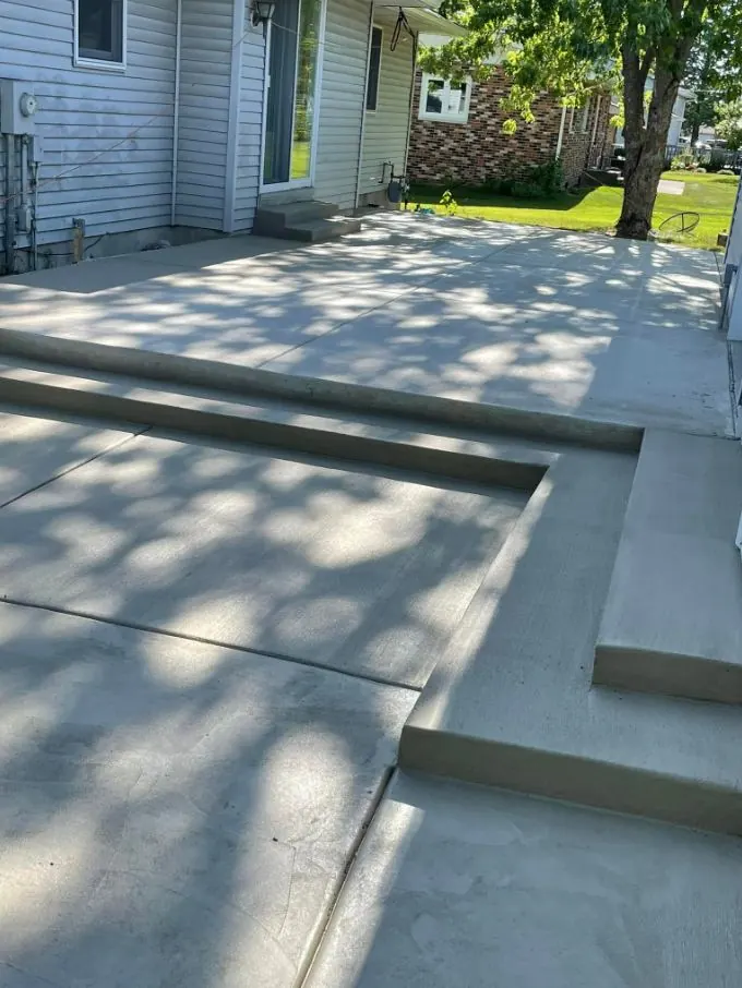 Multi-level broom-finished concrete patio with steps, shaded by trees behind a house