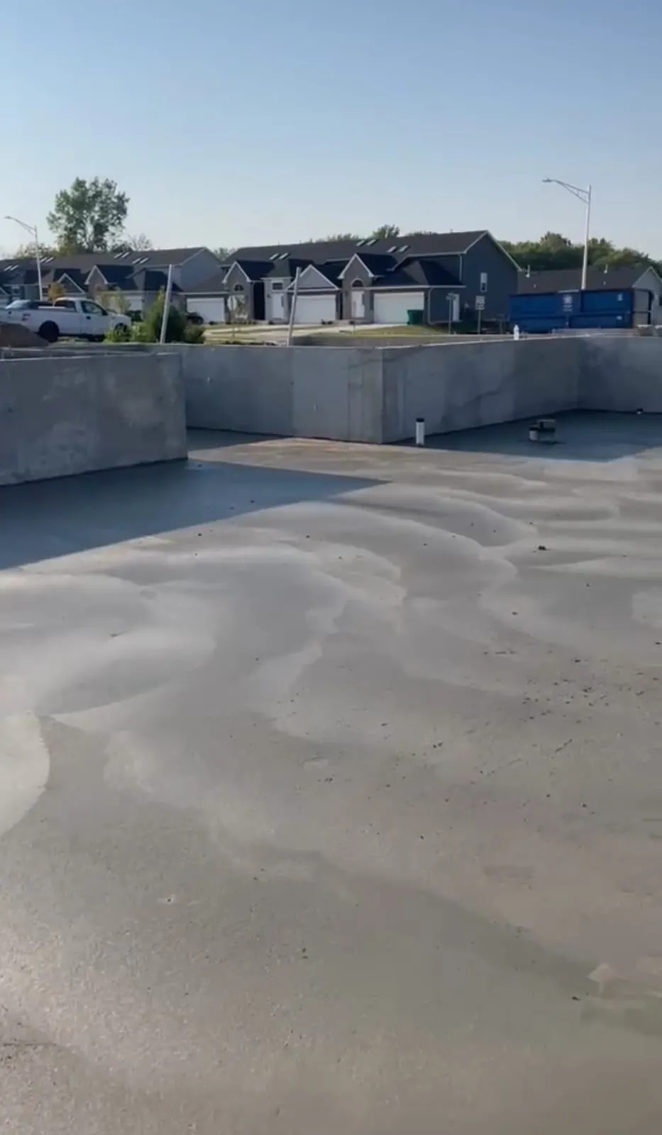 Freshly poured basement slab with wet finish inside foundation walls, new subdivision homes in the background